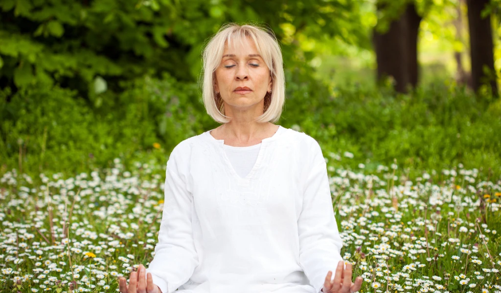 Eine Frau mittleren Alters sitzt in einem wei&szlig;en Outfit auf einer Wiese, umgeben von kleinen wei&szlig;en Blumen. Sie hat die Augen geschlossen und h&auml;lt ihre H&auml;nde in einer Meditationshaltung. Der Hintergrund zeigt &uuml;ppiges Gr&uuml;n, das eine ruhige und friedliche Atmosph&auml;re schafft. Die Frau scheint in tiefer Entspannung und Achtsamkeit vertieft zu sein.