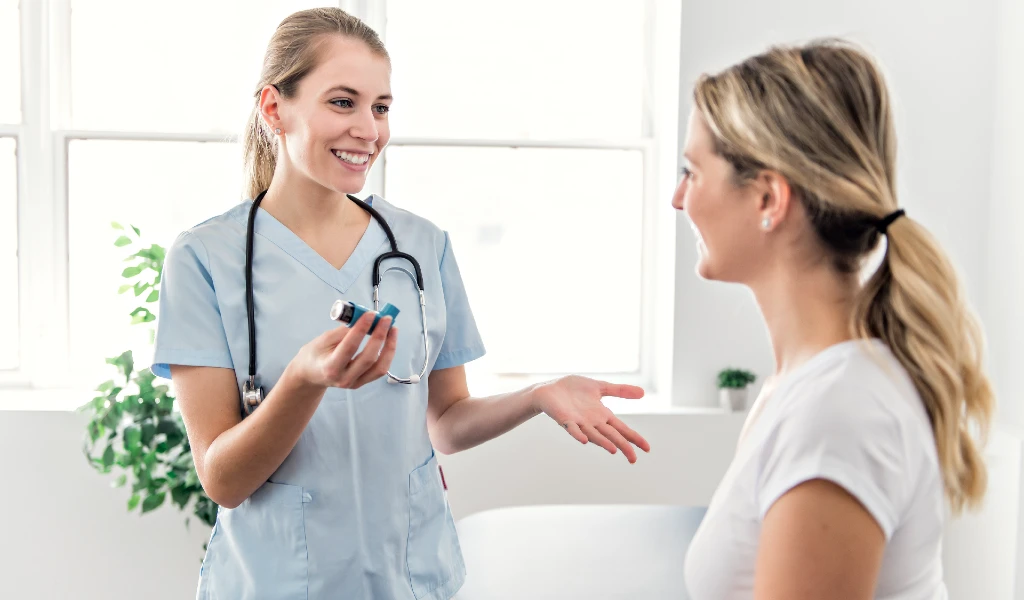 Eine l&auml;chelnde &Auml;rztin h&auml;lt einen Inhalator und erkl&auml;rt einer Patientin dessen Verwendung.