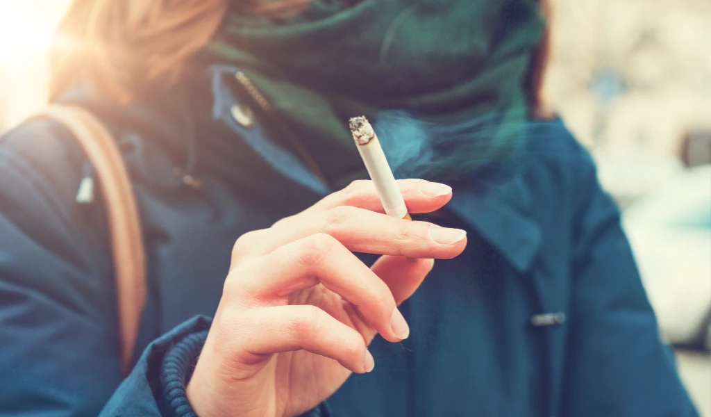Eine Person im Mantel h&auml;lt eine brennende Zigarette in der Hand, in der Luft ist Rauch zu sehen.