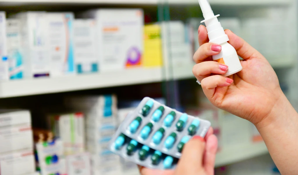 Hand hält Nasenspray und Tablettenschachtel vor Apothekenregal.